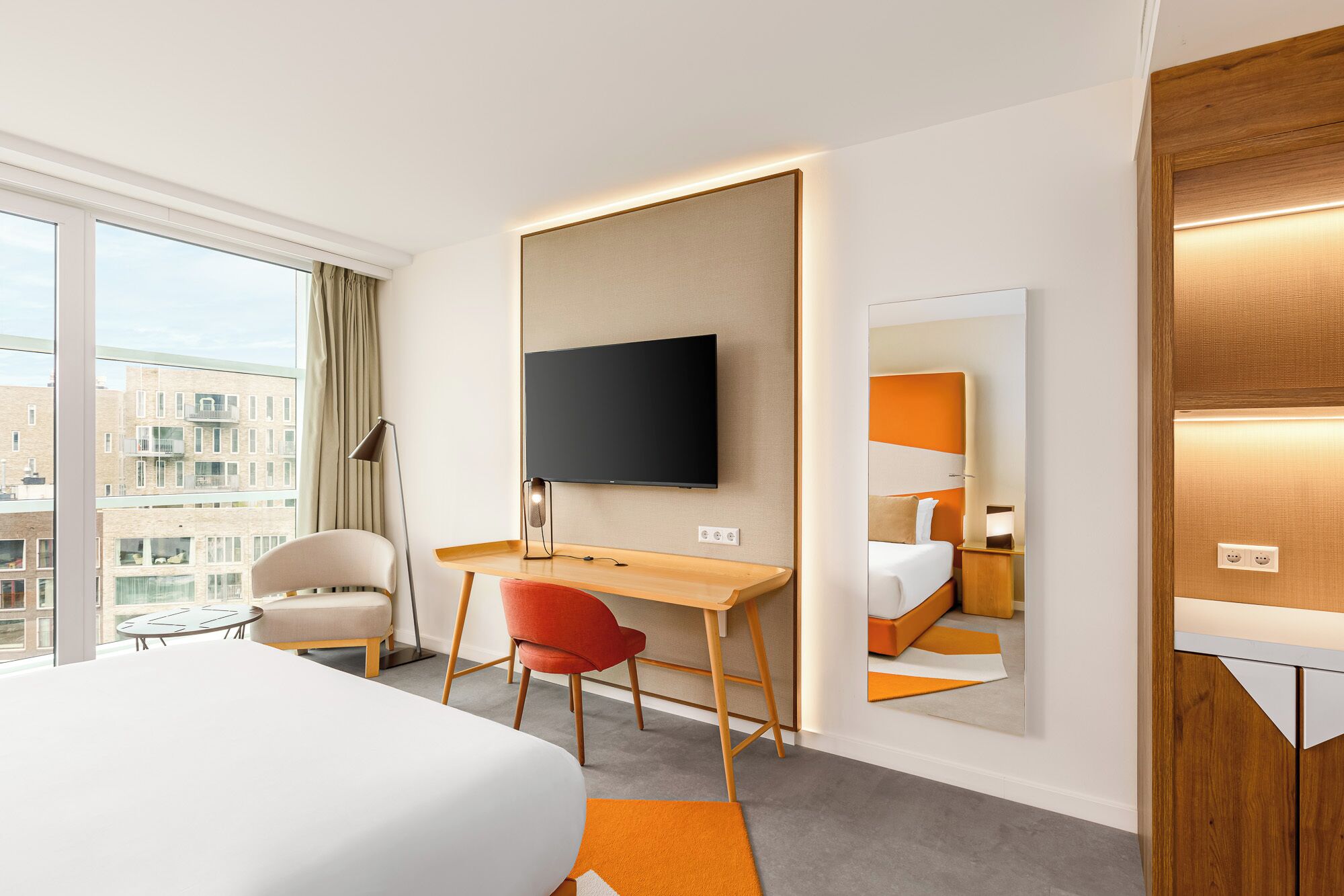 A hotel room with a large bed, a desk with a chair, a television mounted on the wall, and a mirror reflecting the room.