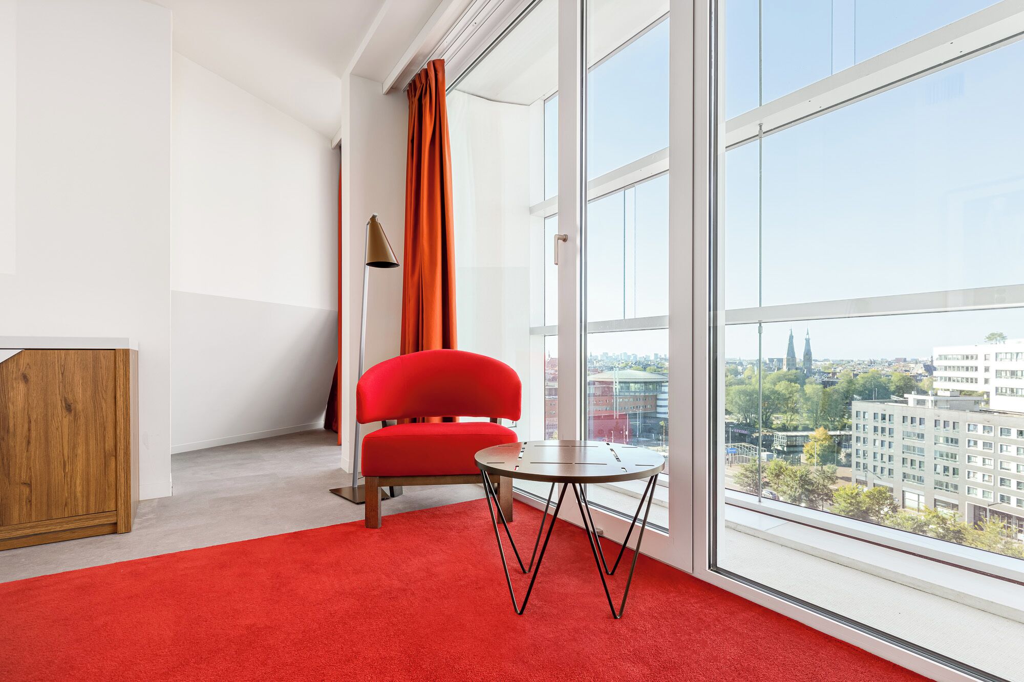 A modern room with a red chair, a small round table, and a large window offering a city view.