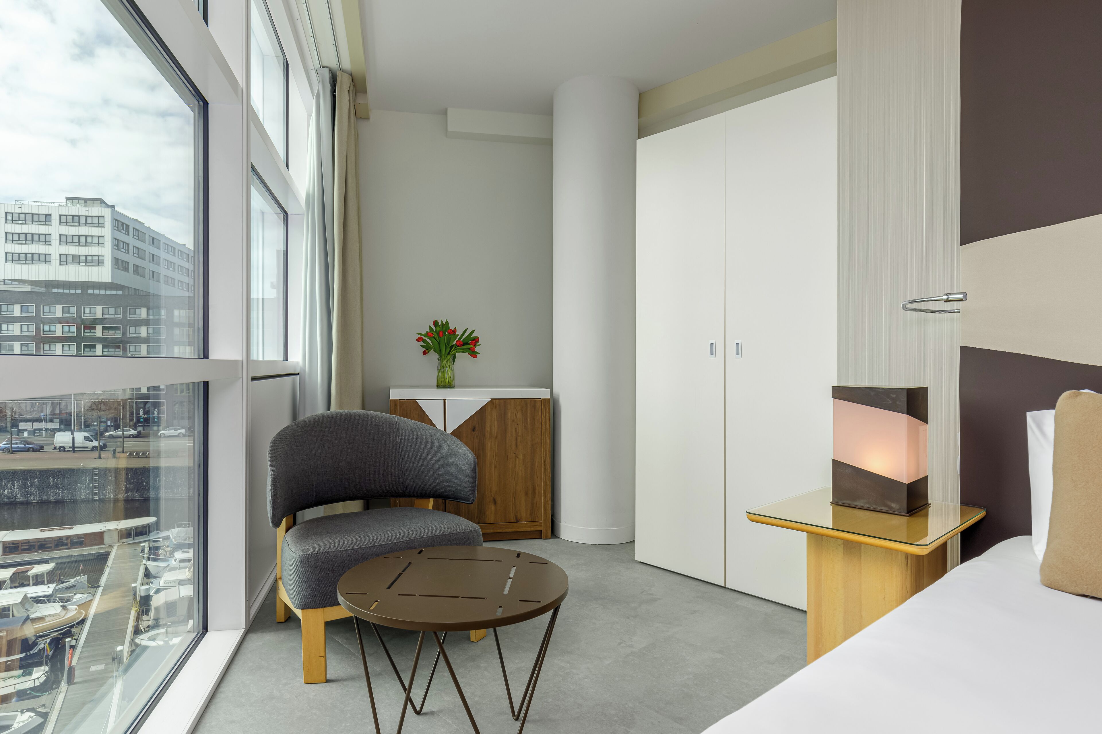 A modern hotel room with a large window, a sofa, a small table, a cabinet, and a bedside table with a lamp.