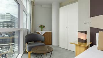 A modern hotel room with a large window, a sofa, a small table, a cabinet, and a bedside table with a lamp.