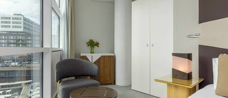 A modern hotel room with a large window, a sofa, a small table, a cabinet, and a bedside table with a lamp.