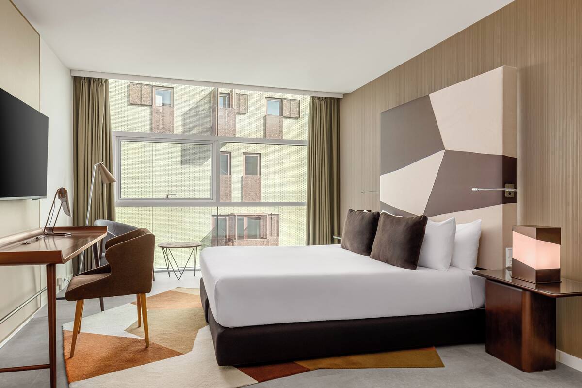 Una habitación de hotel moderna con una cama grande, un escritorio y vista a un edificio a través del ventanal.