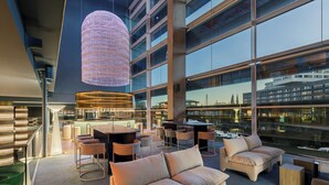 A modern lounge area with seating and tables, large windows offering views of the cityscape, and a distinctive hanging light fixture.