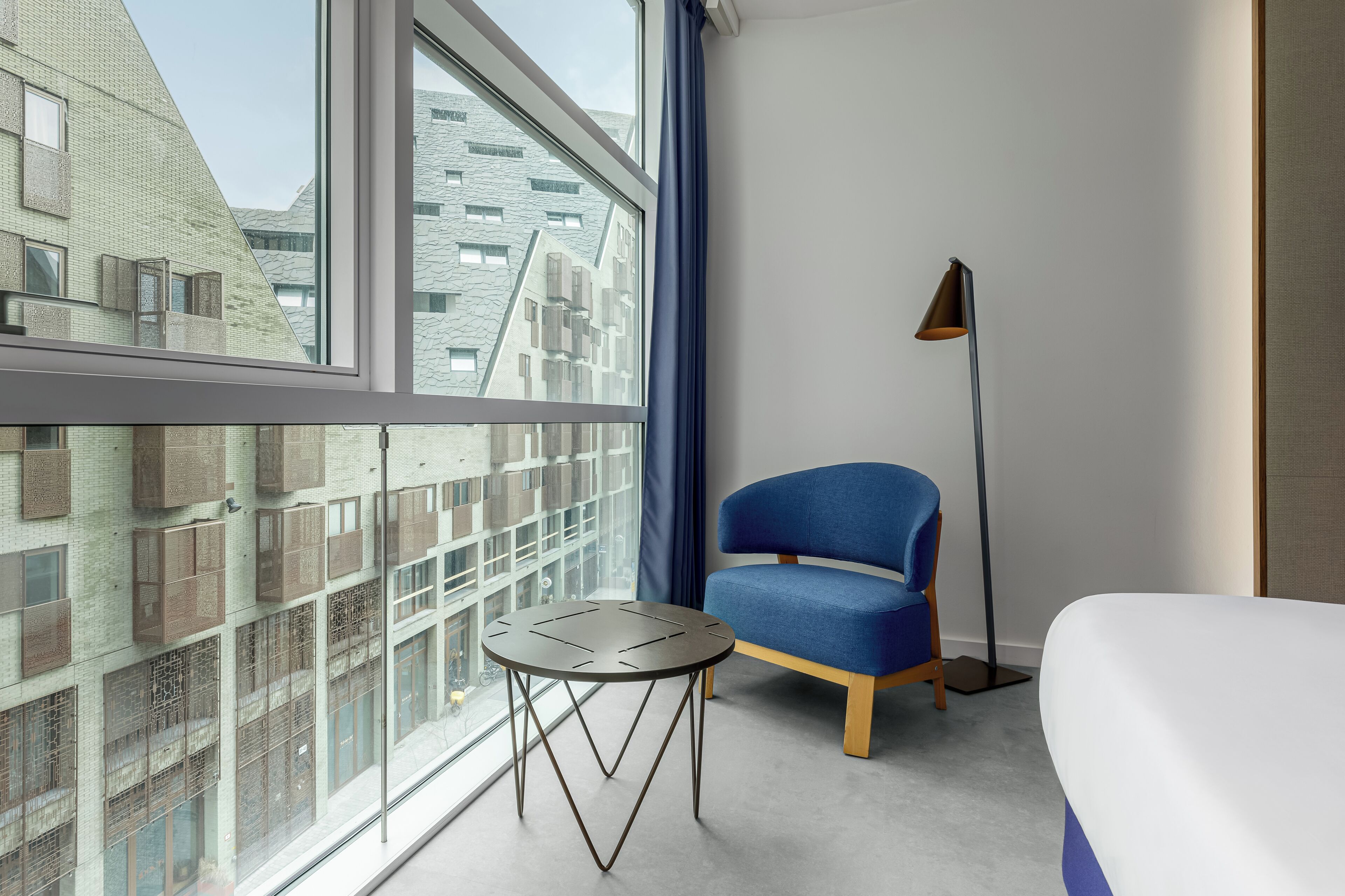 Una habitación de hotel con un ventanal grande, un sillón azul, una mesita redonda y una lámpara de pie.