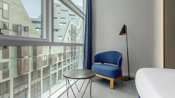 Una habitación de hotel con un ventanal grande, un sillón azul, una mesita redonda y una lámpara de pie.