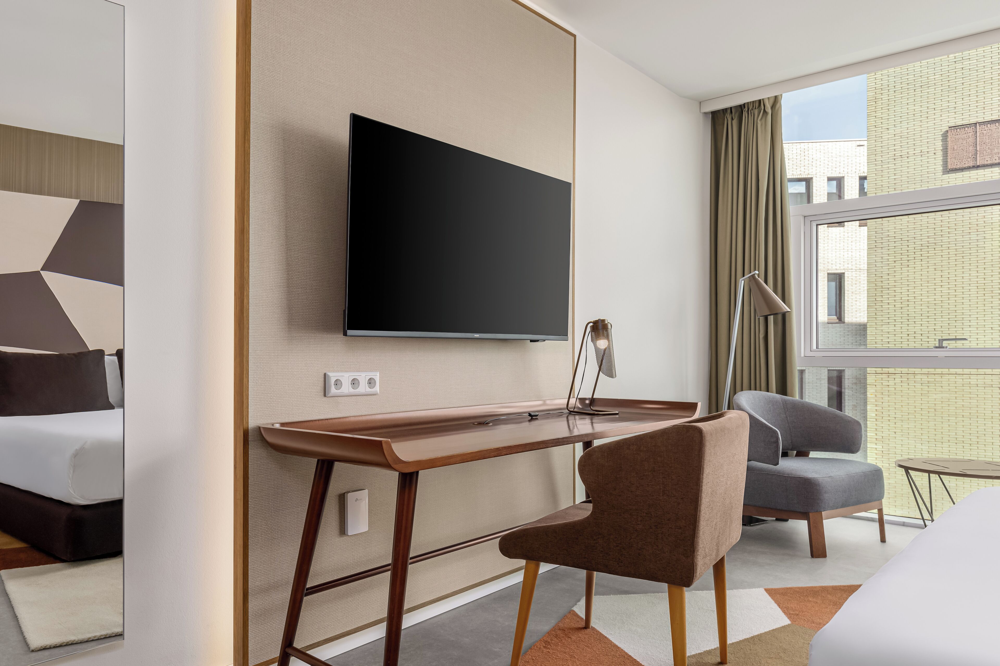 A modern hotel room with a flat-screen TV mounted on the wall, a wooden desk, a grey armchair, and a view of the city through large windows.