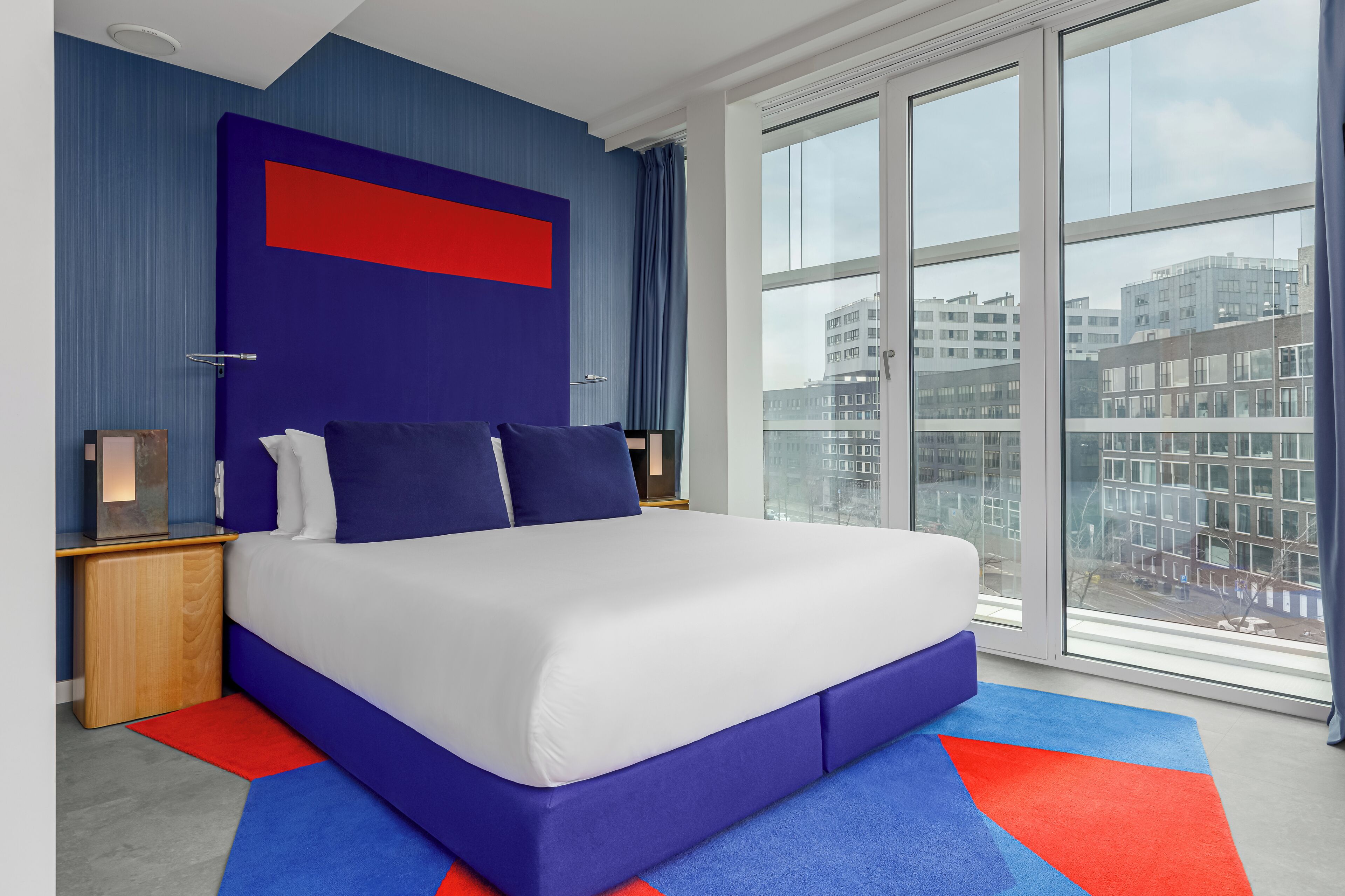 A modern hotel room with a large bed, a city view, and a blue and red accent wall.