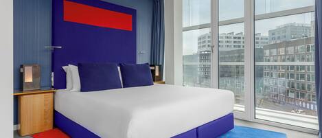 Una habitación de hotel moderna con una cama grande, vistas a la ciudad y una pared decorada en tonos azul y rojo.