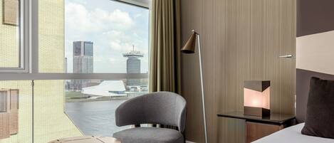 Habitación de hotel moderna con un ventanal grande, vista a la ciudad, una silla, una mesita de noche y una lámpara.