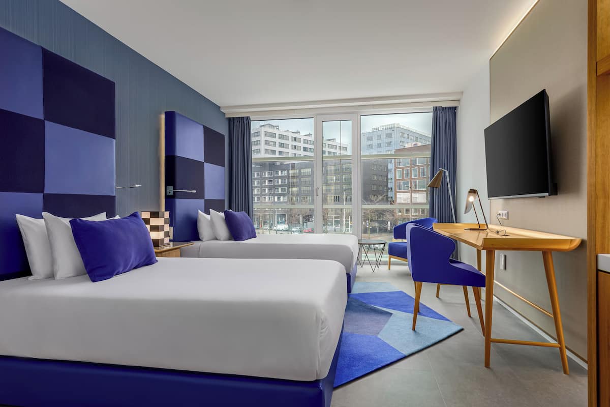 Habitación de hotel moderna con dos camas, un escritorio, una silla, un televisor y ventanas grandes con vistas a la ciudad.