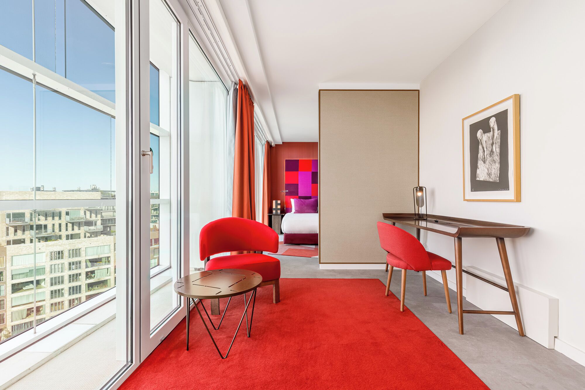 Ein modernes Hotelzimmer mit großem Fenster, roten Stühlen, einem kleinen runden Tisch und einem Bett mit farbenfrohem Kopfteil.