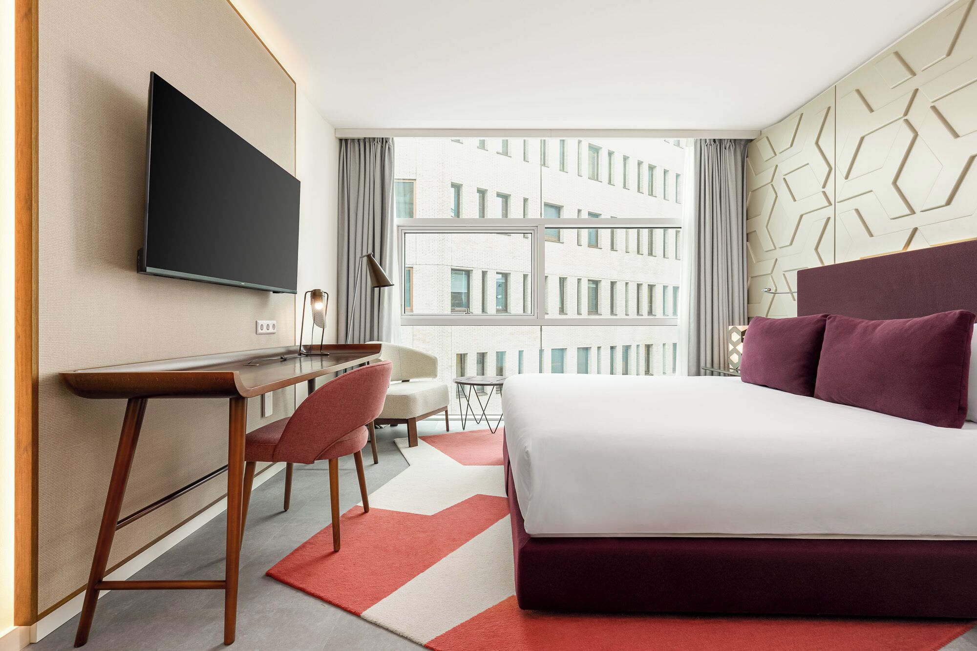 A hotel room with a bed, a desk, a chair, a TV, and a view of buildings.