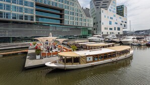 A waterfront area with a modern building, a dock with a boat, and several yachts.