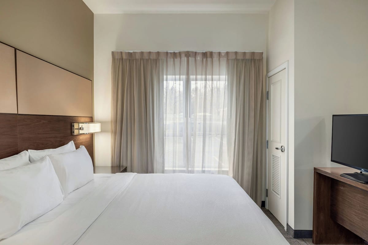 Habitación de hotel con una cama grande, un televisor sobre un mueble de madera, una ventana con cortinas traslúcidas y una puerta con radiador.