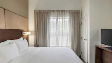 A hotel room with a large bed, a TV on a wooden stand, a window with sheer curtains, and a door with a radiator.