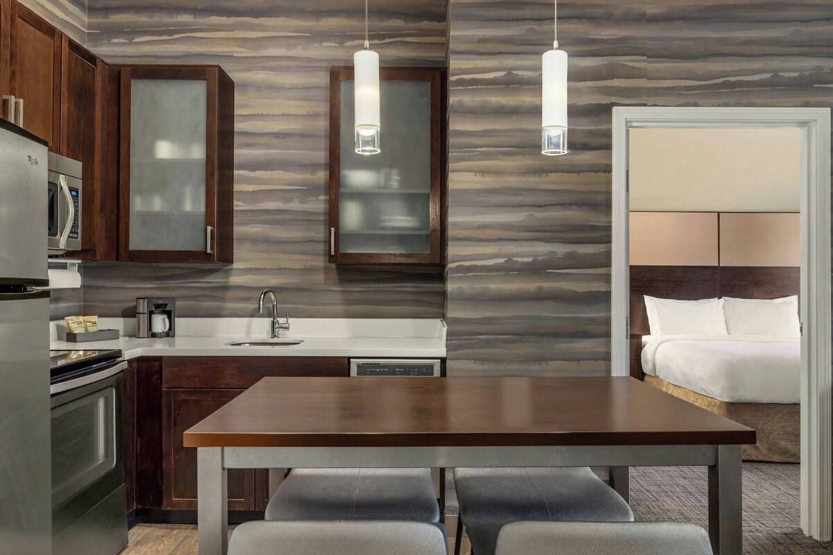 Una cocina moderna con armarios de madera oscura, un refrigerador de acero inoxidable y una encimera blanca. Hay una mesa de comedor con cuatro sillas y lámparas colgantes arriba.