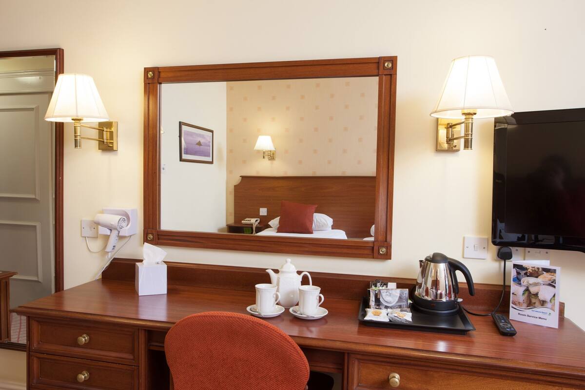 A hotel room with a wooden desk, two wall-mounted lamps, a mirror, a television, a bed with a red pillow, a tea set, and a tray with a remote control and a menu.