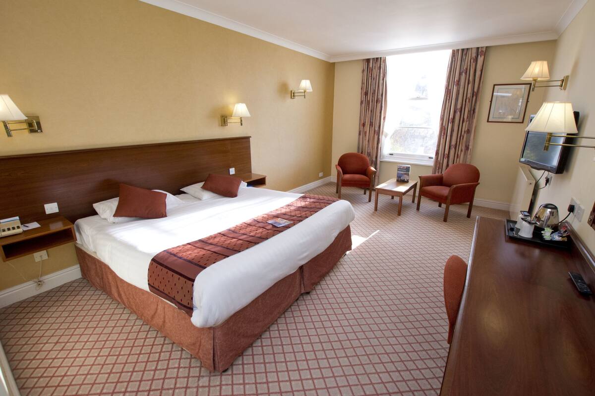 A hotel room with a large bed, two bedside tables, a desk with a chair, a TV, and a window with curtains.