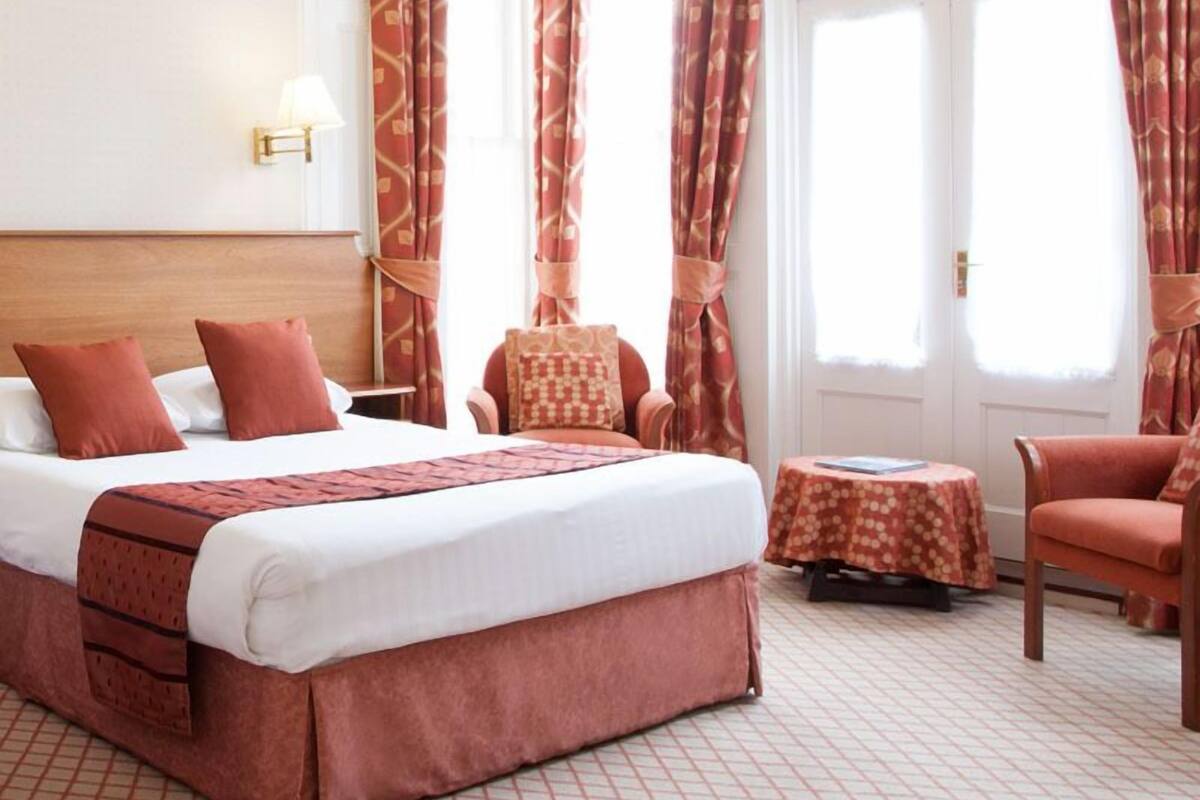 A hotel room with a large bed, two chairs, a small table, and a door with curtains.