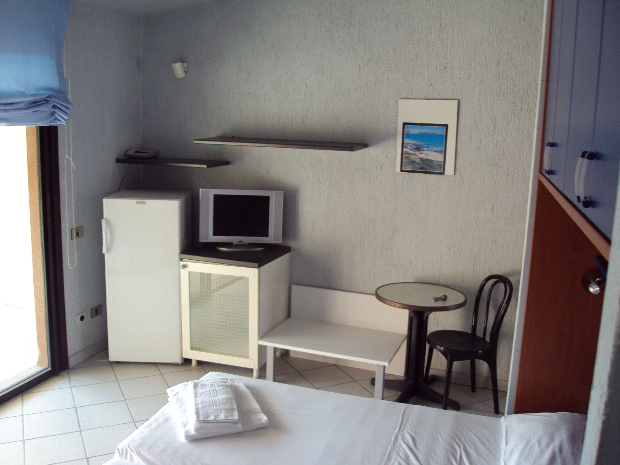 A compact hotel room with a bed, a small desk with a television, a refrigerator, a chair, and a picture on the wall.