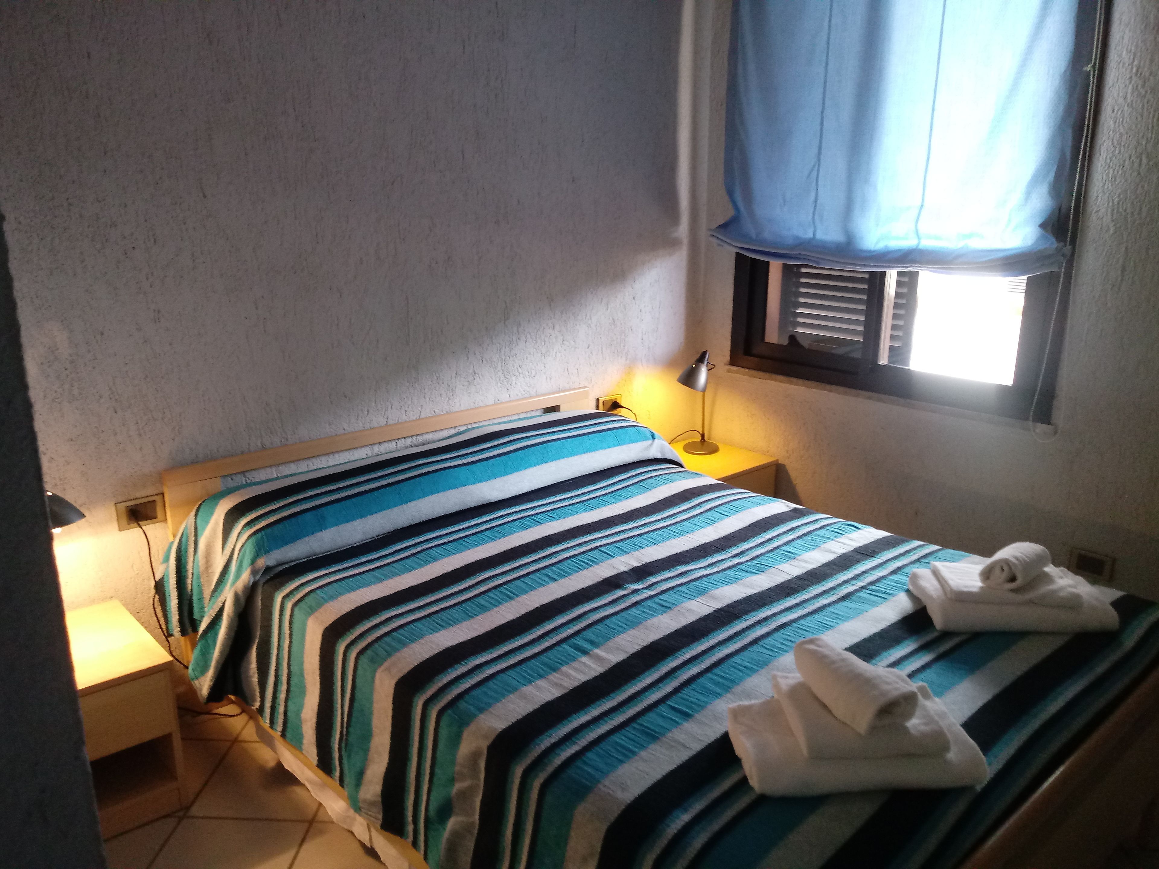 Een kleine, eenvoudige kamer met een bed, een nachtkastje, een lamp en een raam met gordijn.