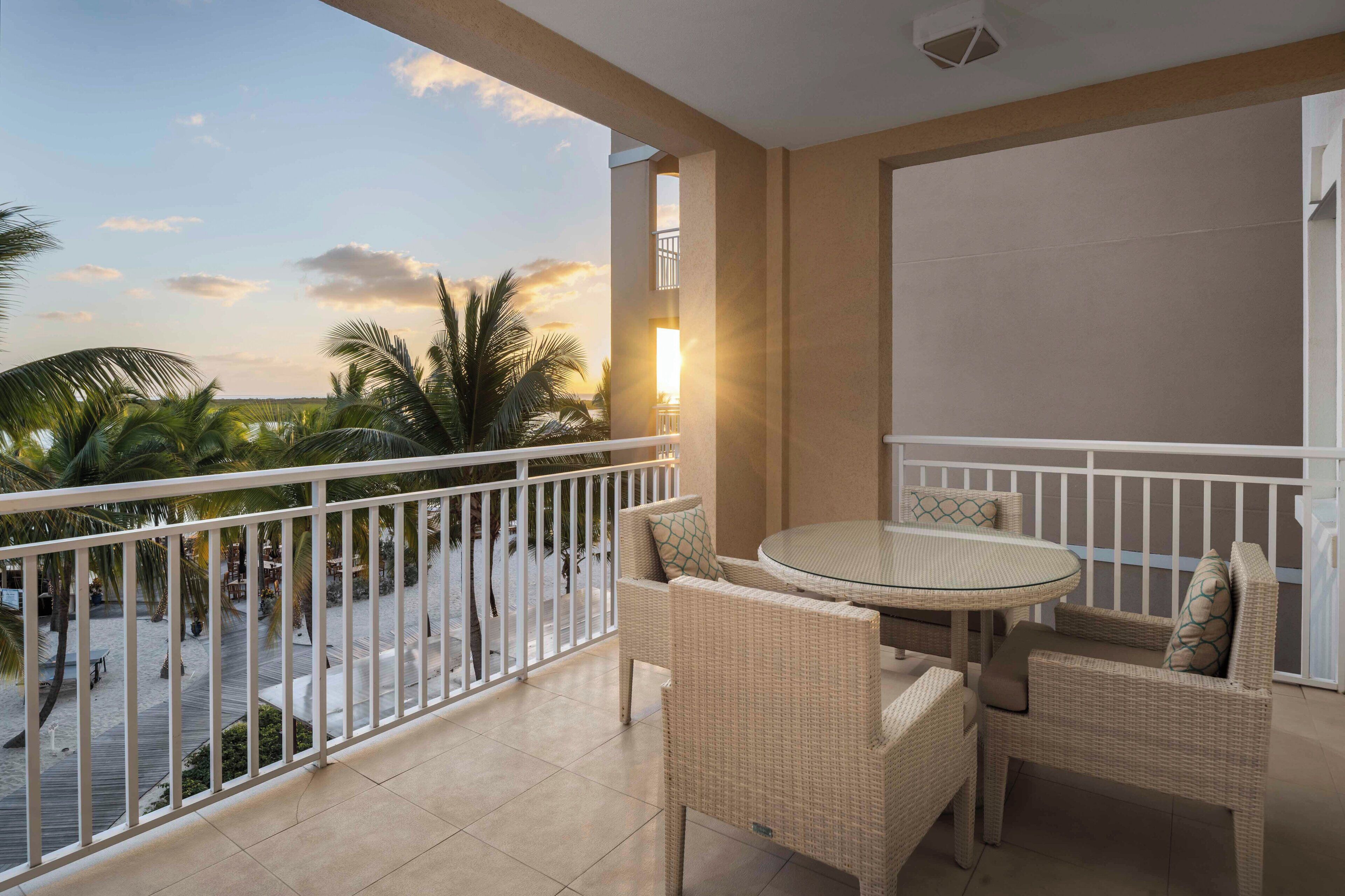 A balcony with wicker furniture, a glass table, and a view of palm trees and a sunset.