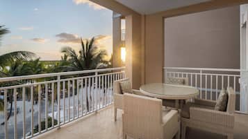 A balcony with wicker furniture, a glass table, and a view of palm trees and a sunset.