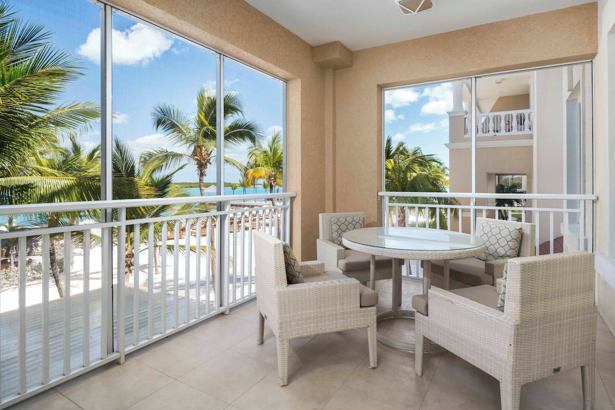 A balcony with a glass railing, wicker furniture, and a view of palm trees and a beach.