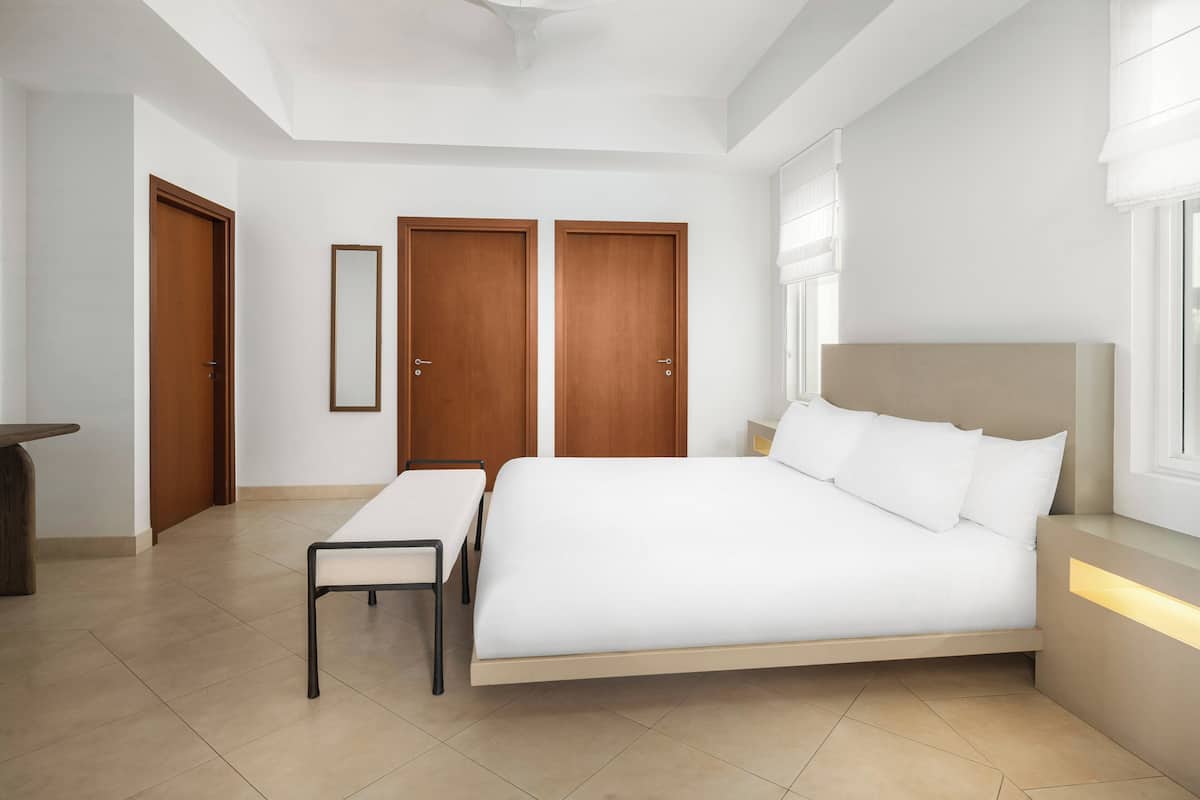 A bedroom with a bed, a bench, two doors, a mirror, and a bedside table.