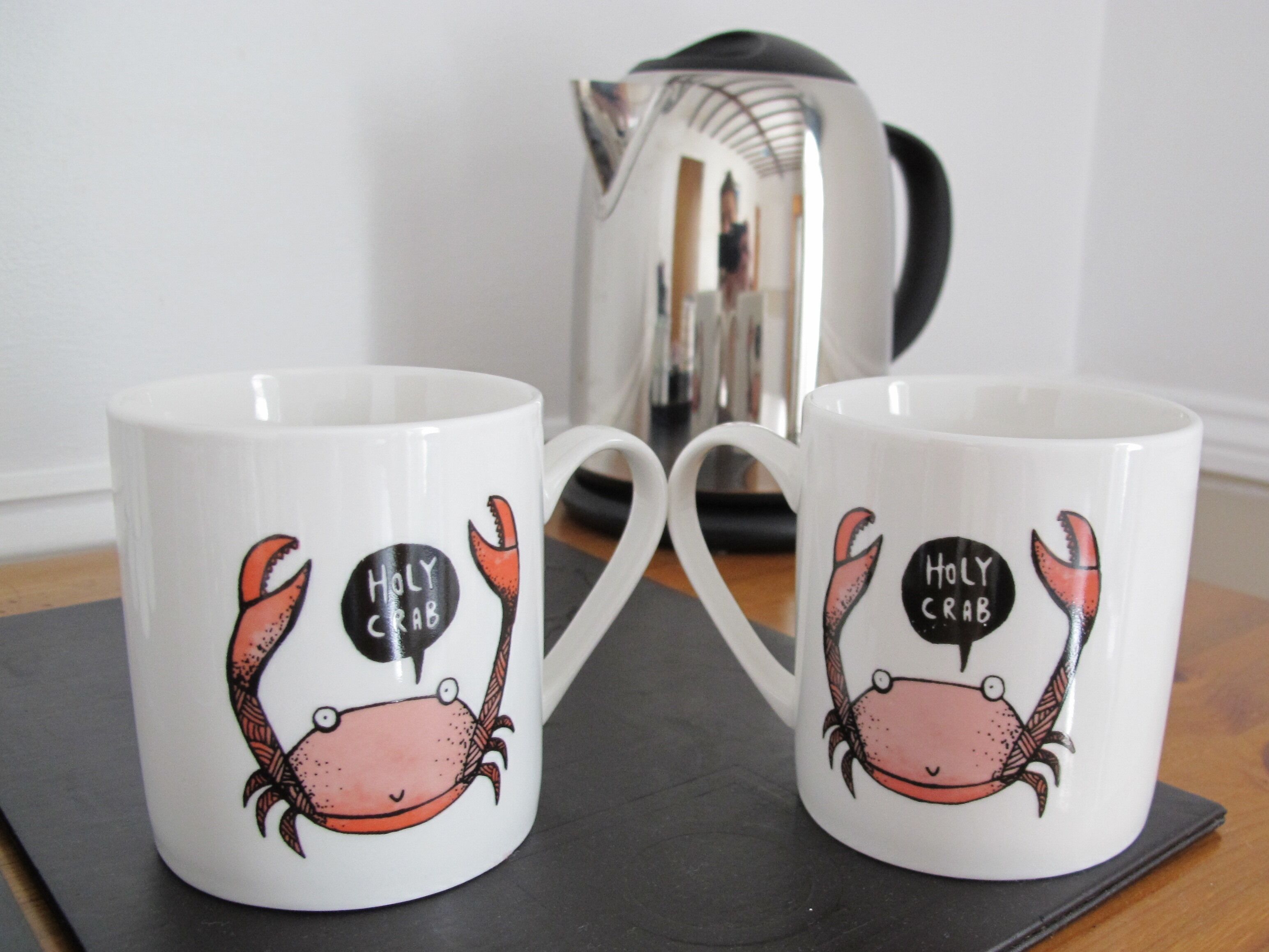 Deux tasses blanches ornées de dessins de crabes et portant l’inscription « HOLY CRAB » sur un fond noir, posées sur un sous-verre noir.