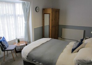 Hotellihuone, jossa on sänky, tuoli, pieni pöytä, puinen vaatekaappi, kello ja ikkuna, jossa on verhot.
