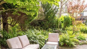Un área de estar al aire libre con dos sillas acolchadas y una mesa de vidrio, rodeada de abundante vegetación y árboles.