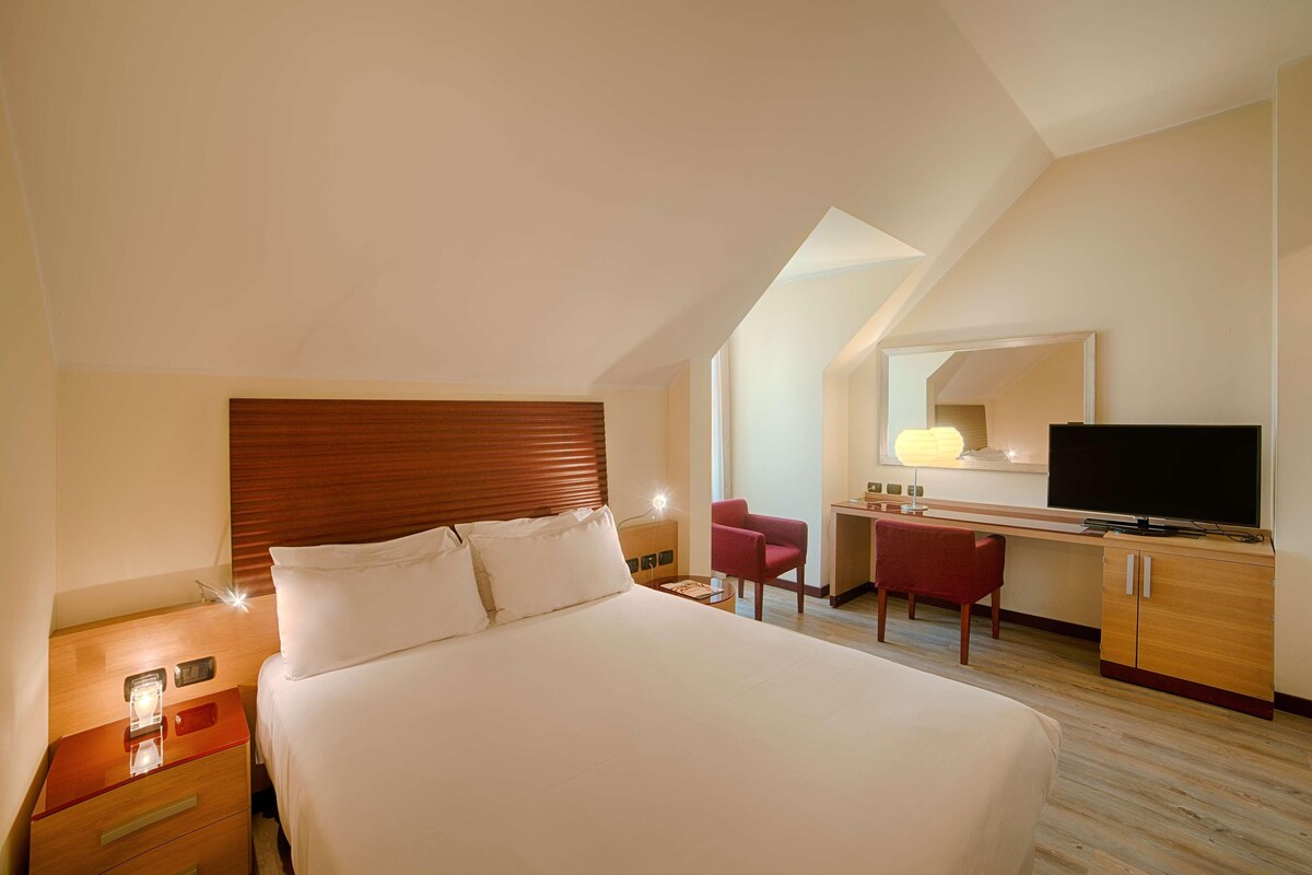 A hotel room with a large bed, a desk with a chair, a television, and a mirror.