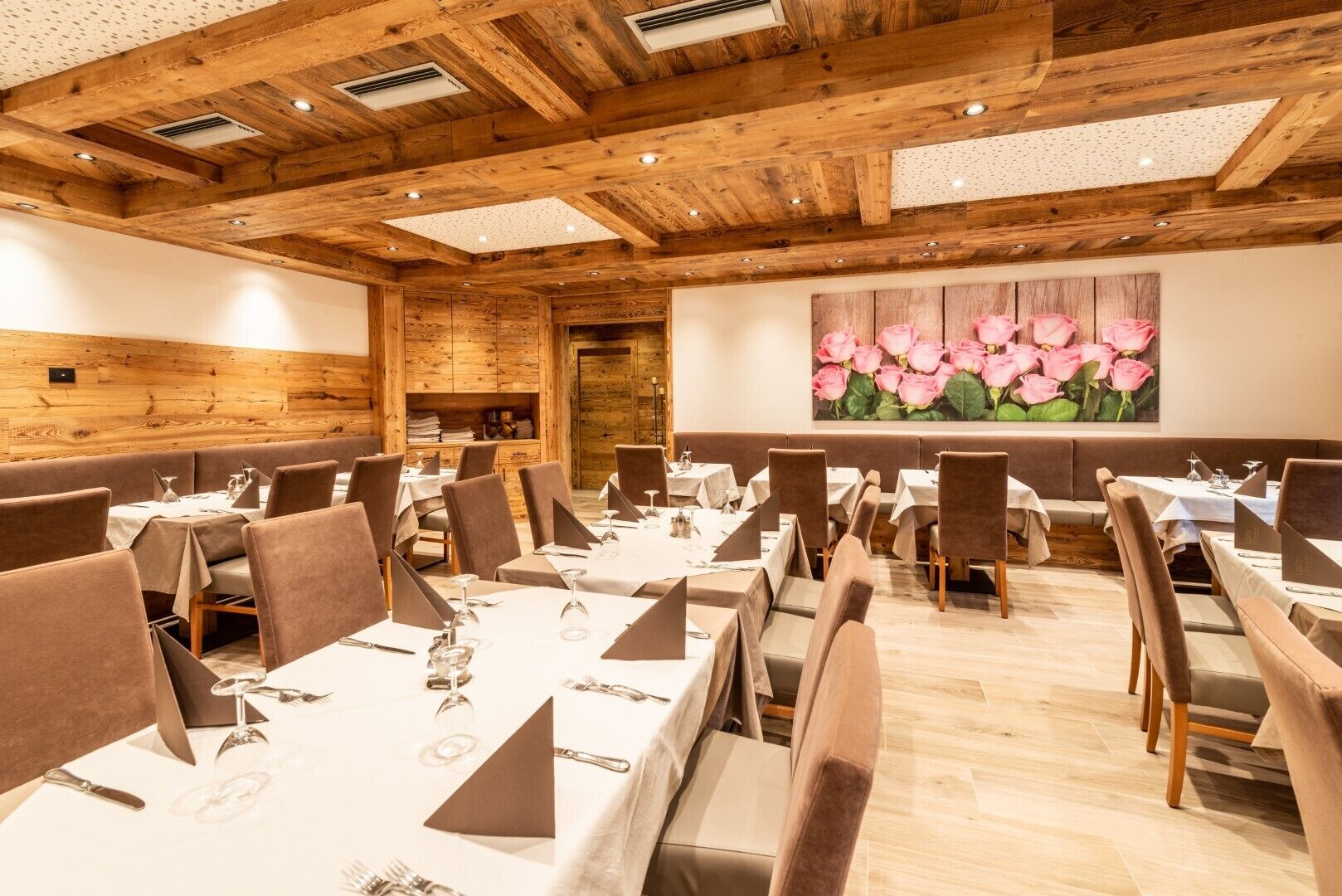 Restaurante com vigas de madeira no teto, mesas brancas com cadeiras marrons e um quadro grande de uma flor rosa na parede.