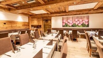 Restaurante com vigas de madeira no teto, mesas brancas com cadeiras marrons e um quadro grande de uma flor rosa na parede.