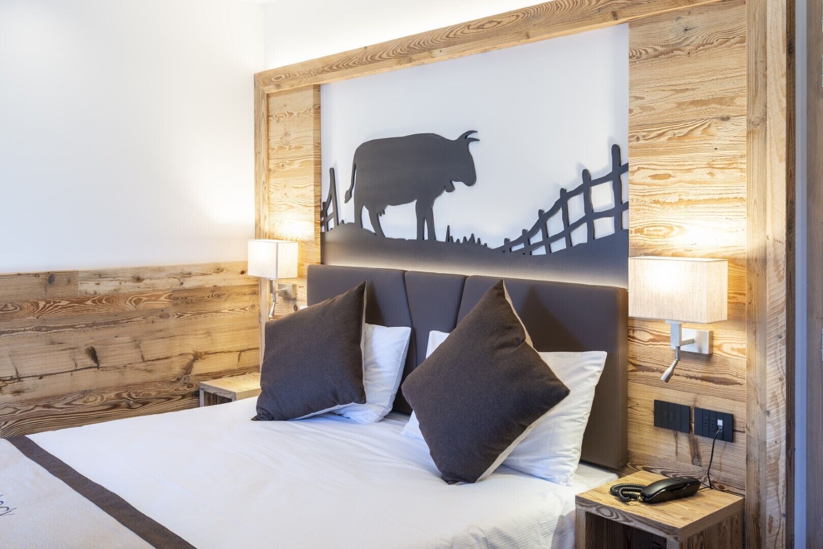 Una camera d'albergo con una testiera in legno, un letto con lenzuola bianche, due cuscini, un comodino con una lampada e un'opera d'arte a parete raffigurante una mucca.