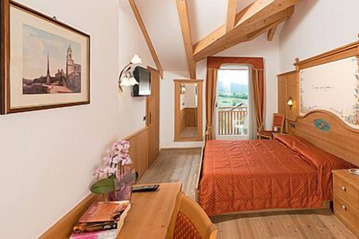 A bedroom with a bed, wooden wardrobe, and a desk with a chair. There is a window with a view of the outdoors.
