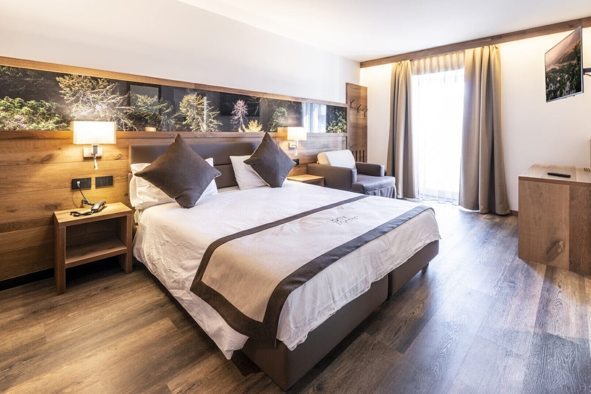 A modern hotel room with a large bed, wooden headboard, bedside lamps, a nightstand, a desk, and a view of trees through a window.