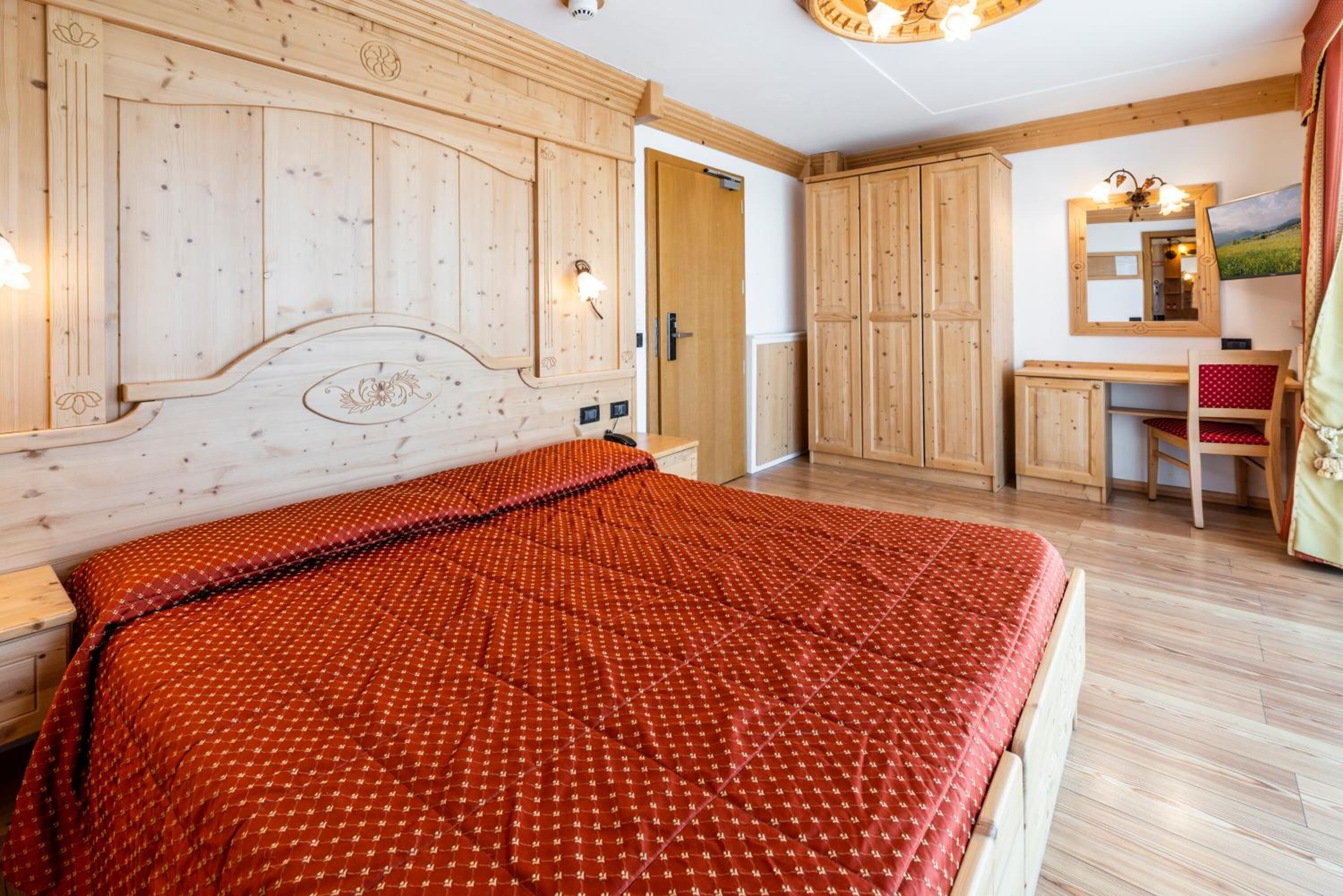 Une chambre avec une tête de lit en bois, un lit recouvert d’une courtepointe à motifs rouges, un bureau avec une chaise et un miroir accroché au mur.