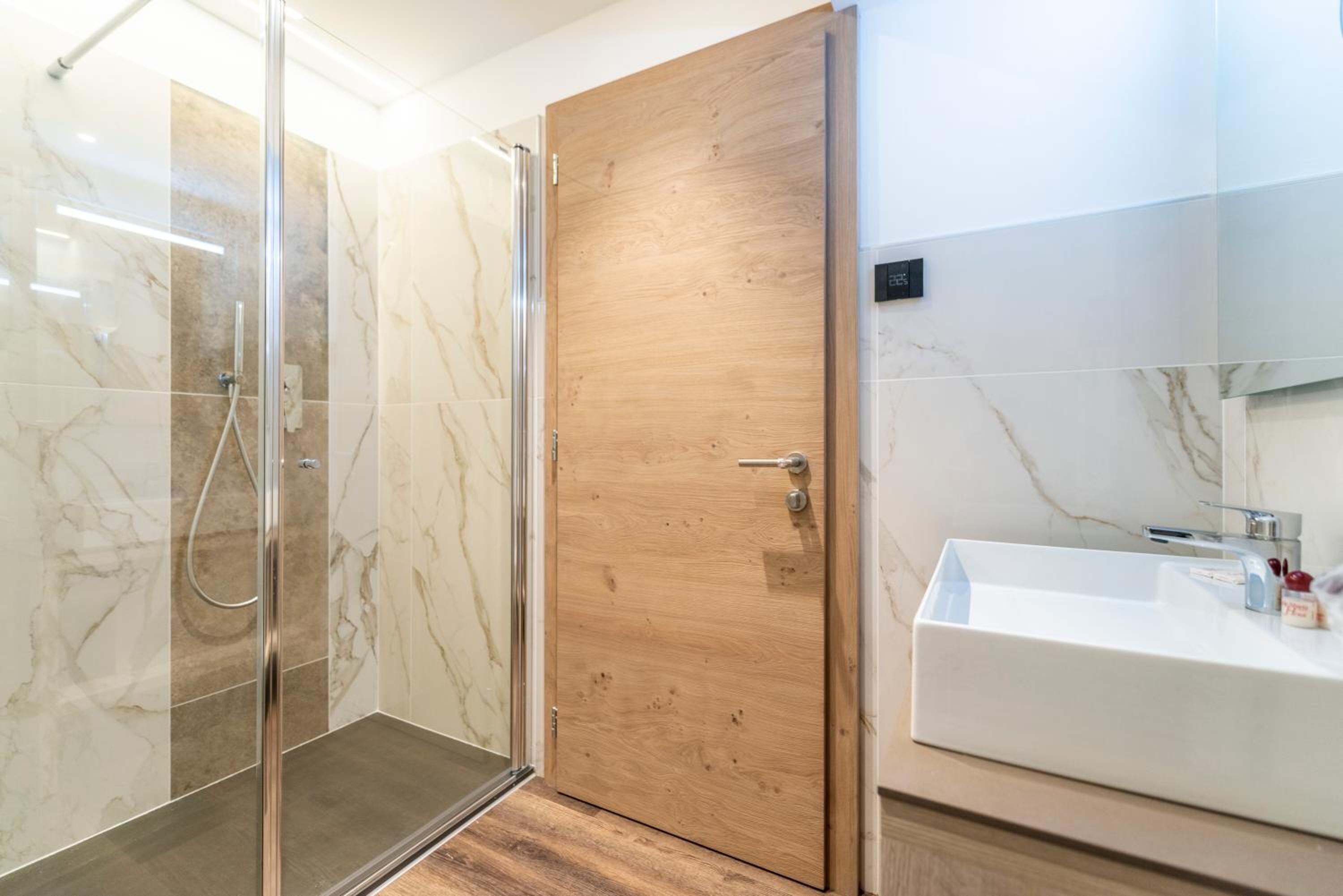 A modern bathroom with a glass-enclosed shower, a wooden door, and a white sink with a mirror.
