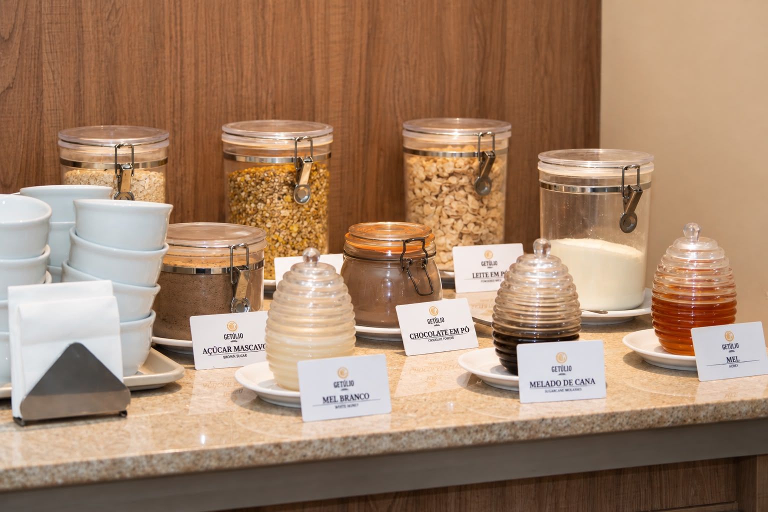 Mostrador de buffet con diversos alimentos en recipientes de vidrio, cada uno etiquetado con el nombre del plato.