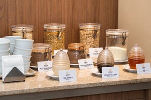 A buffet counter with various food items in glass containers, each labeled with the name of the dish. - Getúllio Hotel (Cuiabá)