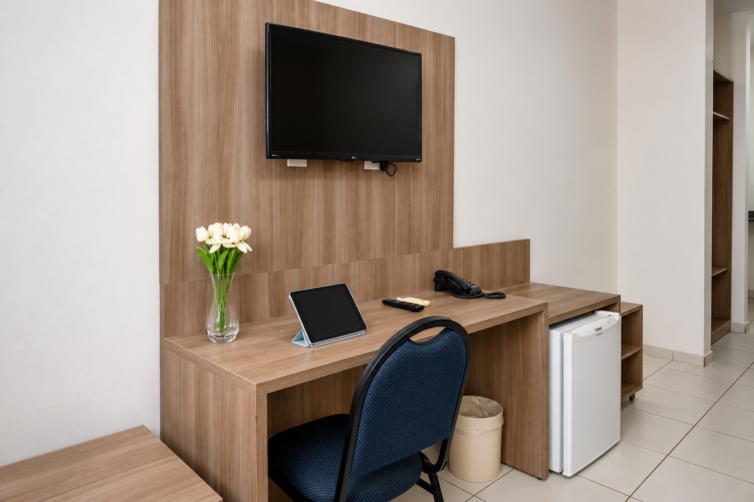 Habitación de hotel con un escritorio de madera, un televisor de pantalla plana montado en la pared, una tableta, un teléfono, un jarrón con flores, una silla y un mini refrigerador.