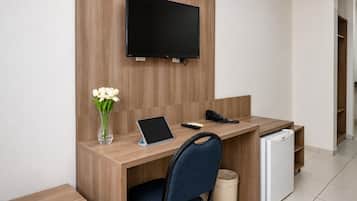 Habitación de hotel con un escritorio de madera, un televisor de pantalla plana montado en la pared, una tableta, un teléfono, un jarrón con flores, una silla y un mini refrigerador.