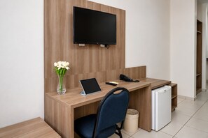 A hotel room with a wooden desk, a flat-screen TV mounted on the wall, a tablet, a telephone, a vase with flowers, a chair, and a mini-fridge. - Getúllio Hotel (Cuiabá)