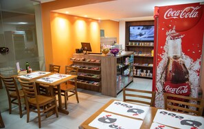A restaurant with wooden tables and chairs, a Coca-Cola vending machine, and a TV screen in the background. - Getúllio Hotel (Cuiabá)