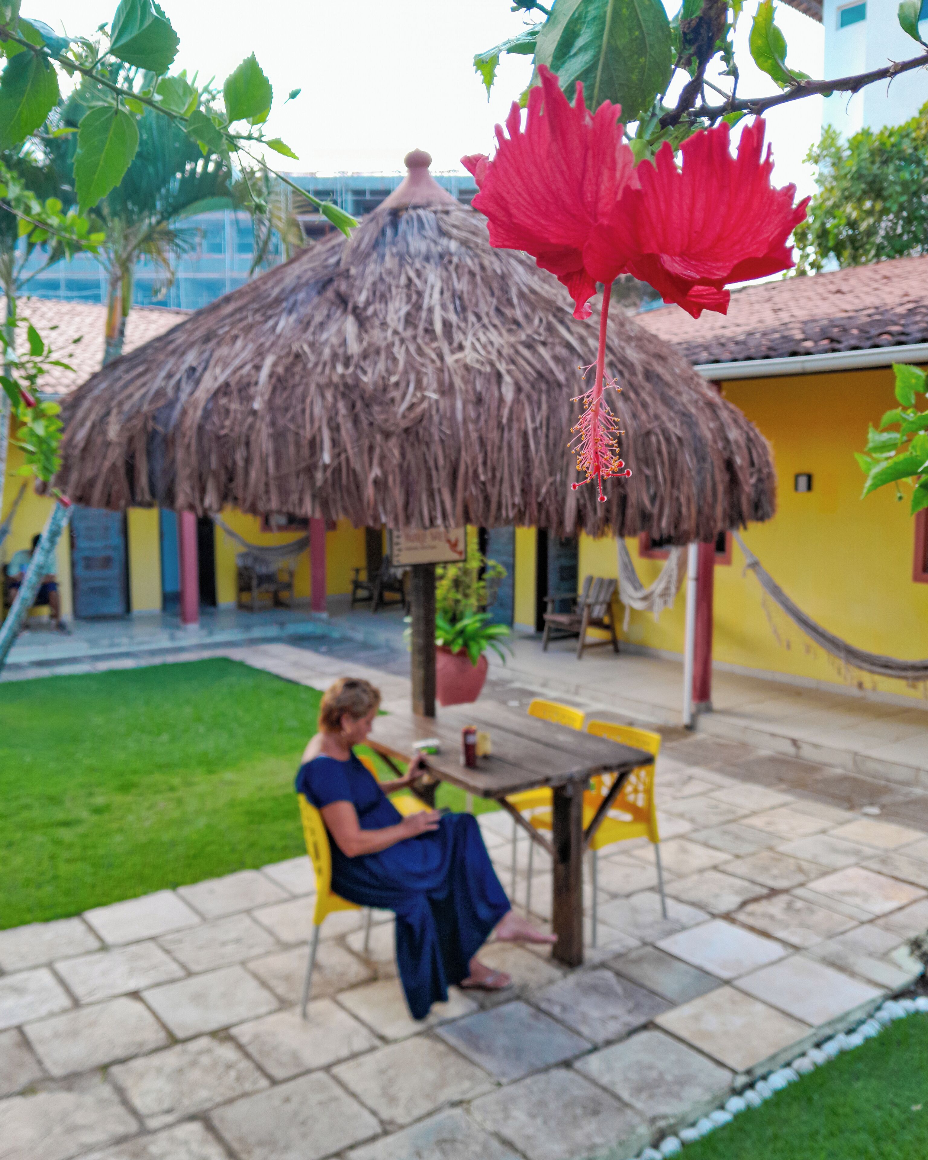 A person sitting at a table under a thatched umbrella in a courtyard with a yellow building in the background.