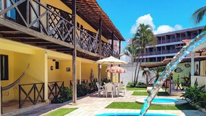 A swimming pool with a clear blue water feature, a yellow building with a balcony, and a palm tree in the background. - Pousada Bore Koko (Ipojuca)