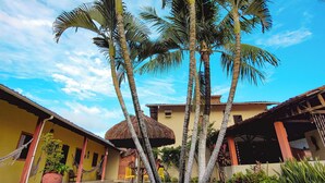 A tropical garden with palm trees, a thatched-roof structure, and a yellow building with a tiled roof. - Pousada Bore Koko (Ipojuca)