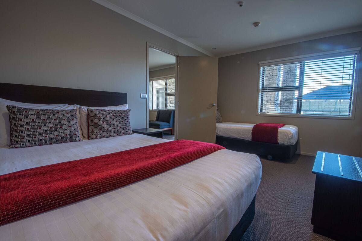 Une chambre d’hôtel avec un grand lit, un lit plus petit, un bureau et une fenêtre avec des stores.